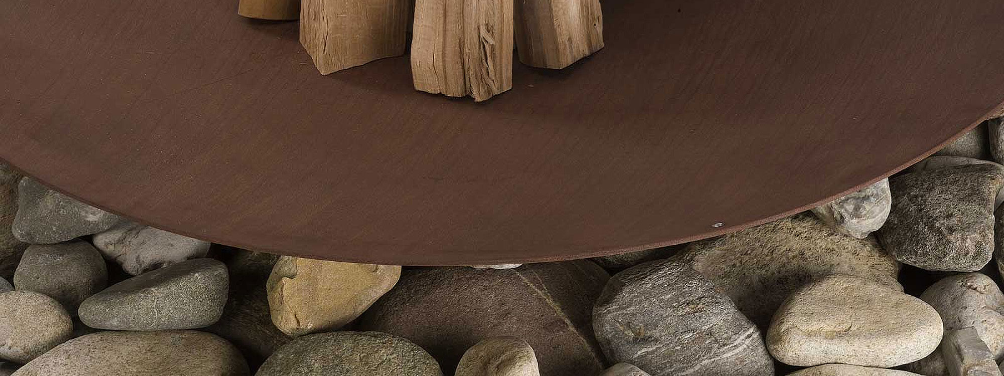 Image of Discolo corten fire bowl by AK47 stacked with logs sat upon large stones