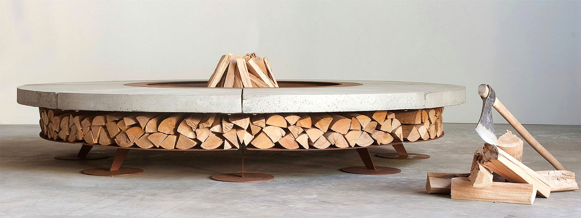Studio image of AK47 Ercole fire pit with integrated log store, with axe and firewood to the side on the floor