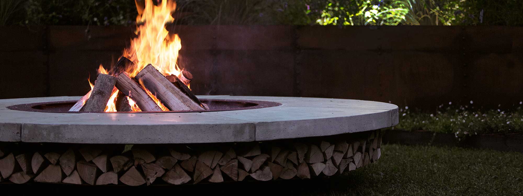 Image of AK47 Ercole large fire pit with natural grey concrete surround, with logs stacked around the fire pit’s circumference.