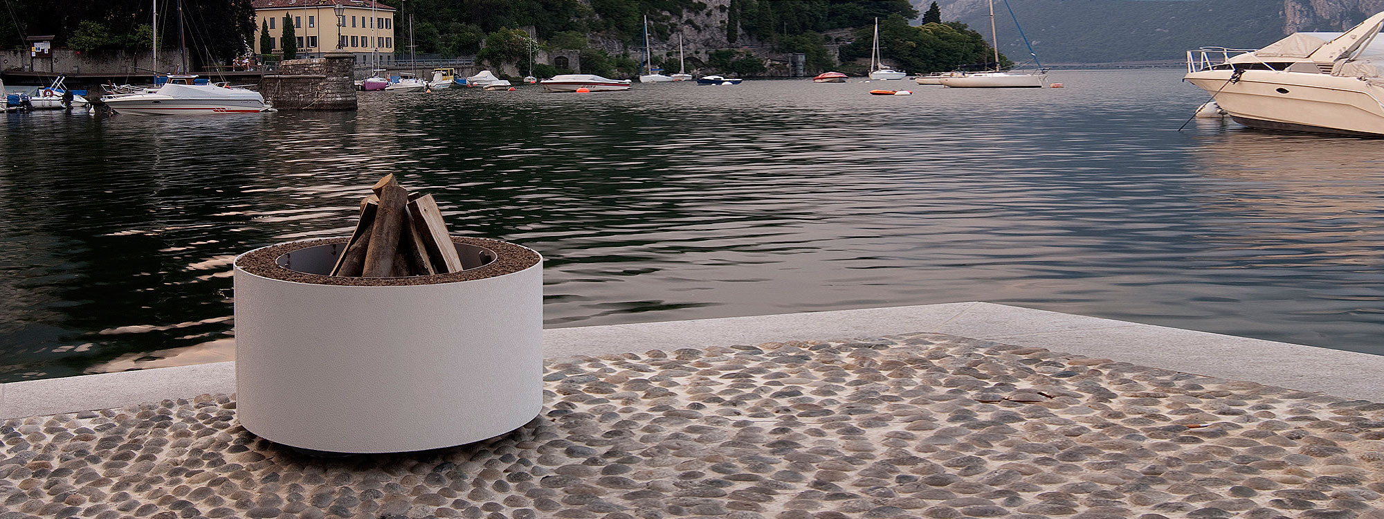 Image of white Mangiafuoco circular fire pit by AK47 with tranquil waters of Lake Como in the background