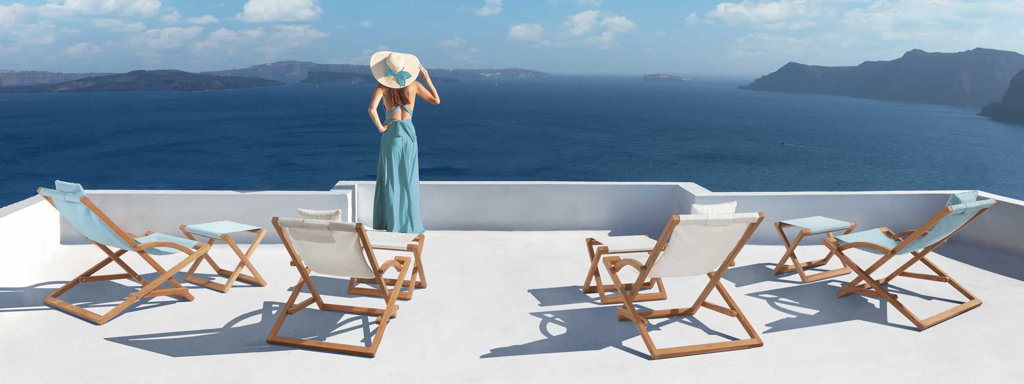 Image of woman looking at Greek islands, surrounded by Beacher deck chairs and foot stools by Royal Botania, with white and blue seat slings.