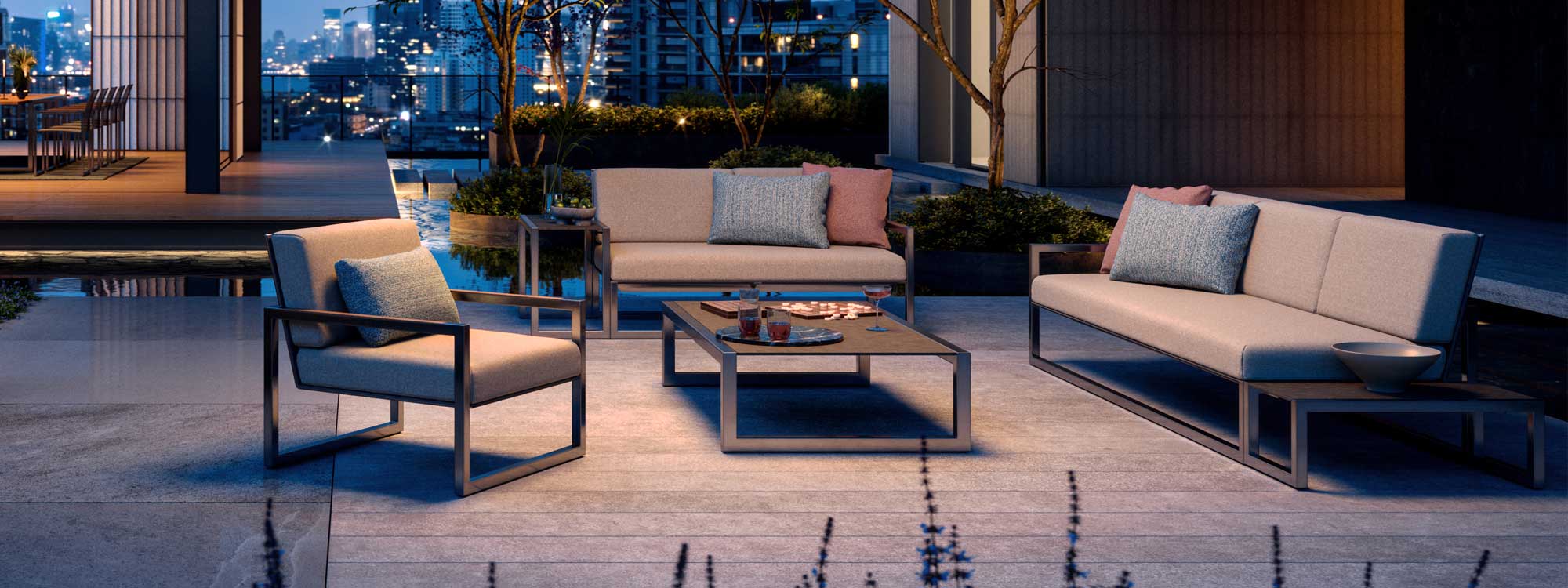 Nighttime image of Royal Botania Ninix Lounge 2 seat garden sofa, 3 seat garden sofa and lounge chairs on rooftop terrace, with city lights twinkling in the background