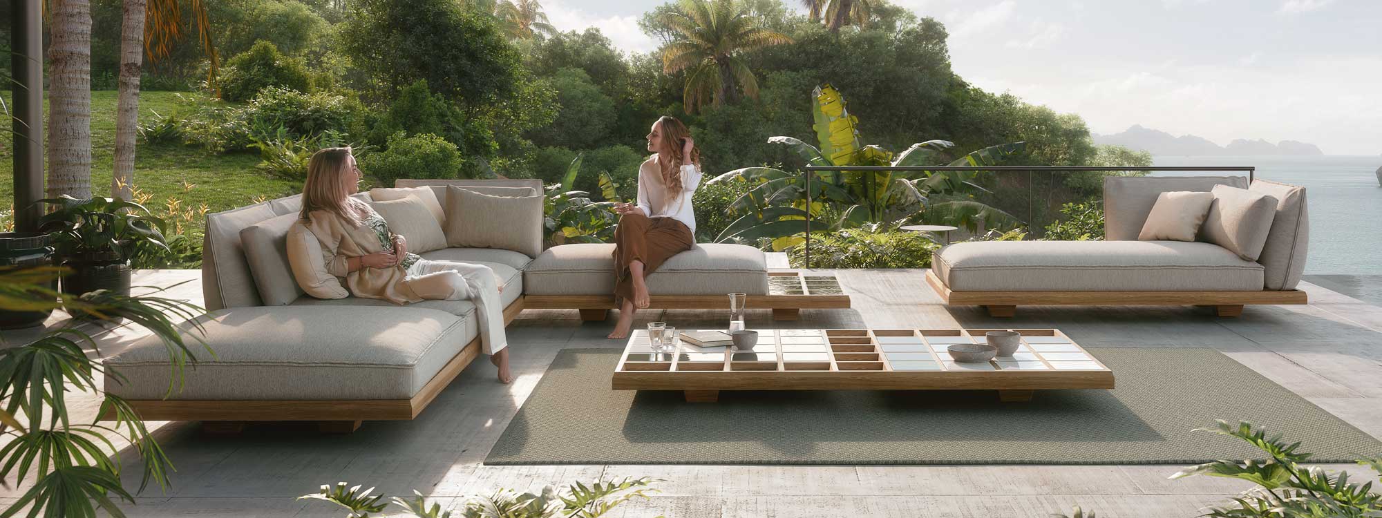 Image of pair of women relaxing on Mozaix modern teak corner sofa and daybed by Royal Botania, with tropical planting and coastline in the background