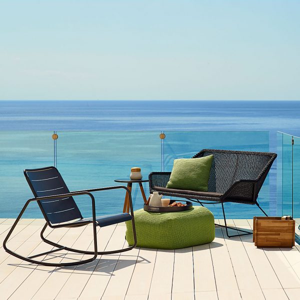 Image of black Copenhagen outdoor rocking chair and black Breeze garden sofa with green Divine pouf on decked outdoor terrace with sea in the background