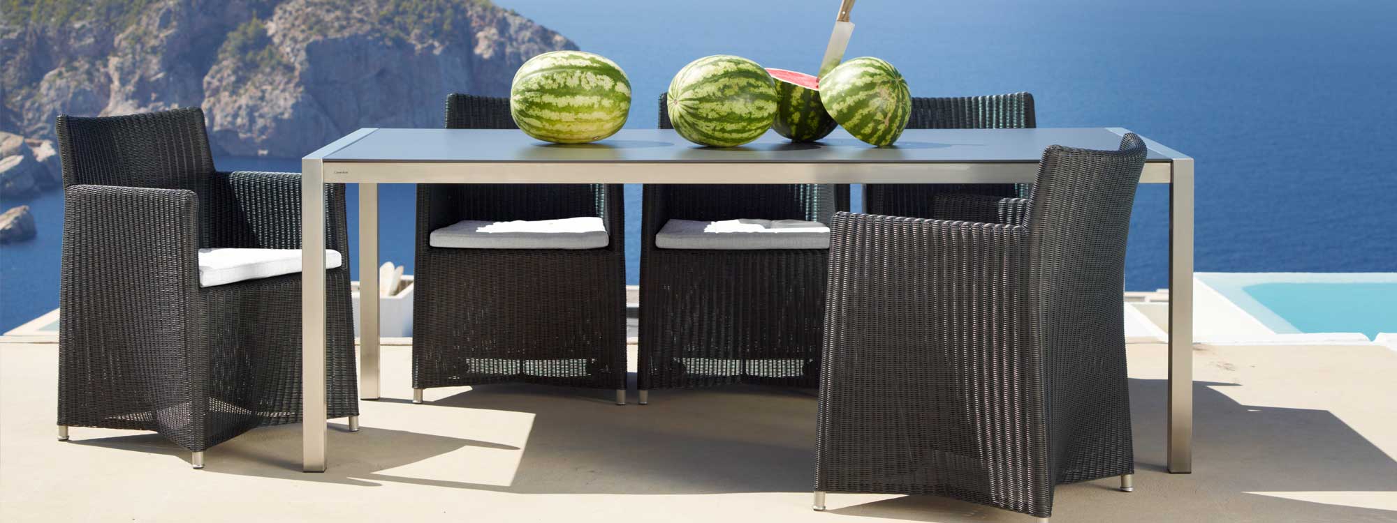 Image of Diamond black woven garden chairs and Cane-line stainless steel dining table with water melons on table top, with dramatic backdrop of sea and headland