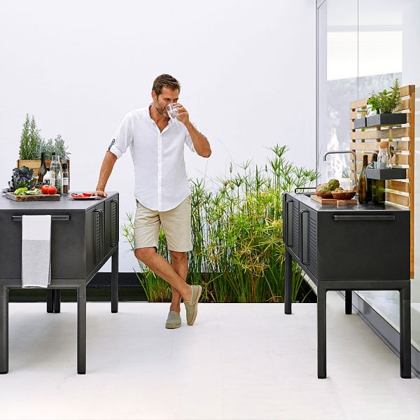 Image of man drinking glass of water, whilst leaning on Cane-line Drop outdoor kitchen island in lava-grey aluminium with Fossil black ceramic top