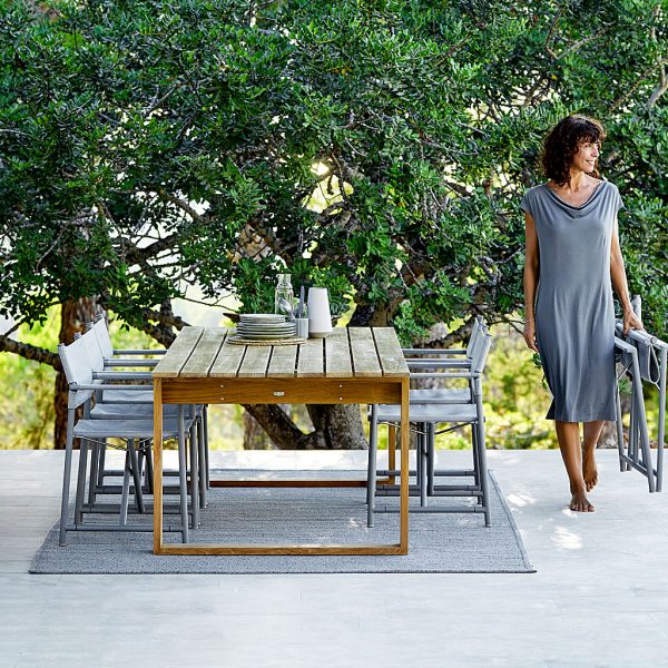Image of woman walking next to Cane-line Endless teak table on terrace, with trees in background