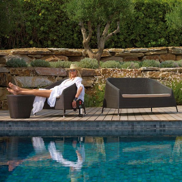 Image of woman on poolside sat on Cane-line Kingston outdoor lounge furniture, with stone terrace and olive tree in background