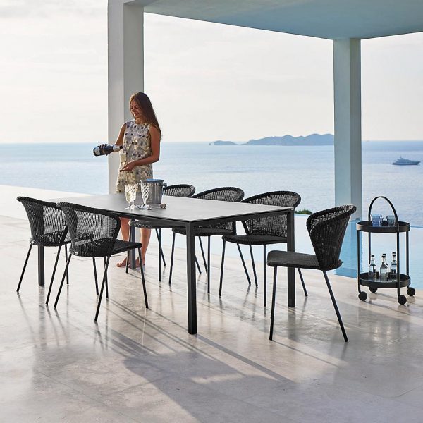 Image of woman stood net to black Lean garden chairs and Drop rectangular garden table by Cane-line, shown on covered seaside terrace