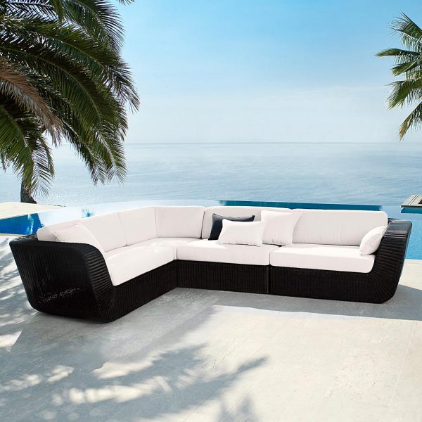 Image of black Savannah corner sofa with white cushions by Cane-line, shown on sunny terrace beneath palm trees with sea in background
