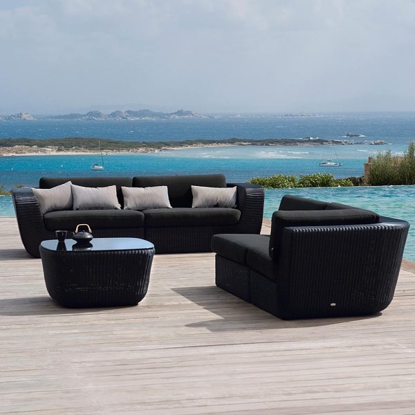 Image of Savannah rattan garden sofa in black weave with black cushions by Caneline, with choppy sea and white horses in background