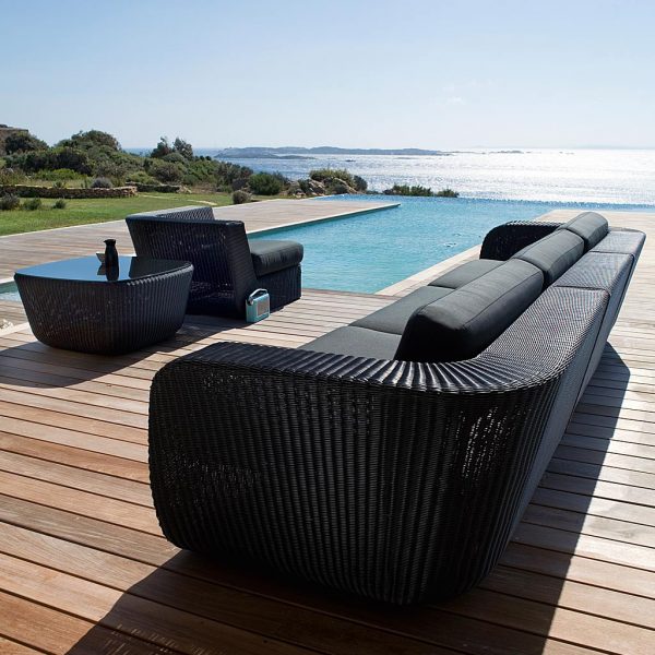Image of Savannah long garden sofa in black weave with black cushions by Cane-line, with shimmering sea in the background