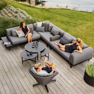 Image of 2 women and a man sat on Caneline Space corner sofa and Peacock swivel lounge chair in dark grey, shown on wooden decking next to lawn and sea