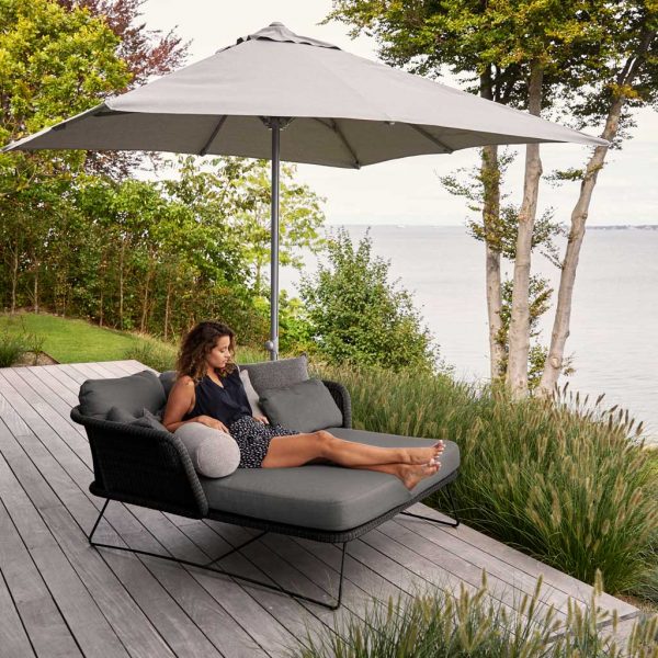 Image of woman relaxing in Horizon garden daybed by Caneline, on decked terrace overlooking water