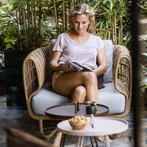 Image of woman sat reading in Caneline Nest outdoor lounge chair in natural Cane-line Weave with Taupe cushions, with lush planting in background