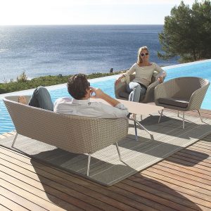 Image of couple sat relaxing on Shell taupe garden sofa and easy chair by FueraDentro, shown on wooden decking next to swimming pool with trees and sea in the background