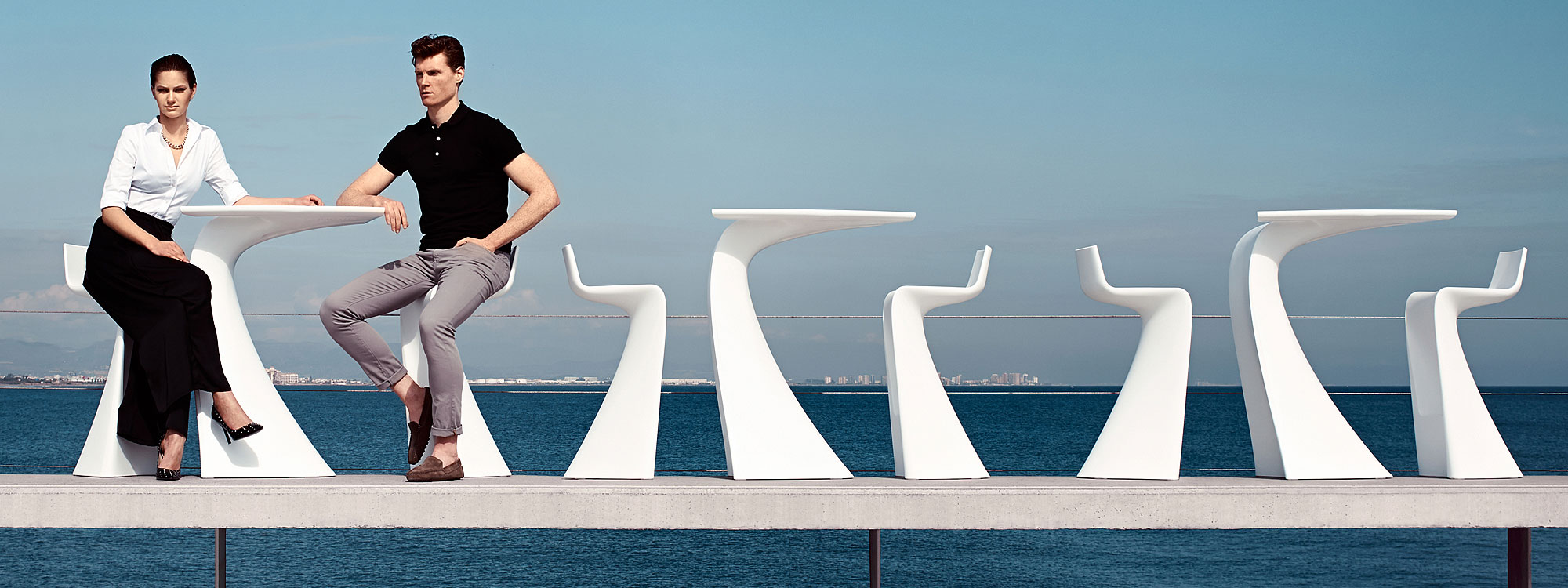 Image of man and woman sat on Vondom Wing contract bar furniture with blue sea and sky in the background