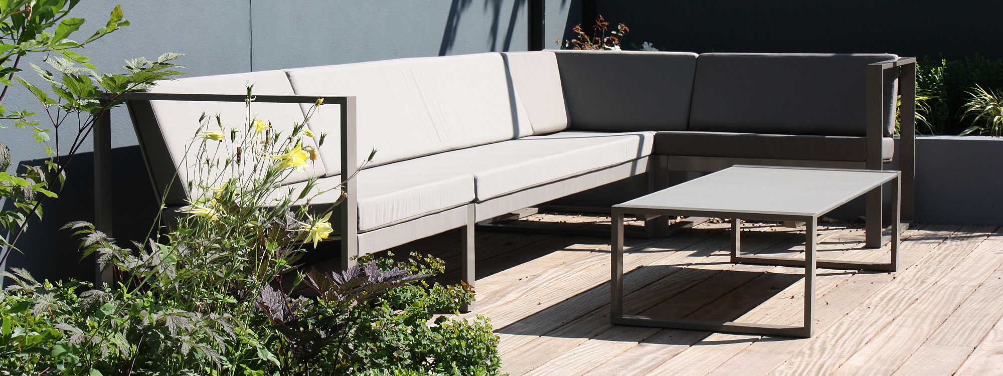 Image of FueraDentro Cima Lounge modern garden corner sofa with taupe frame and taupe cushions, shown on sunny decking in London