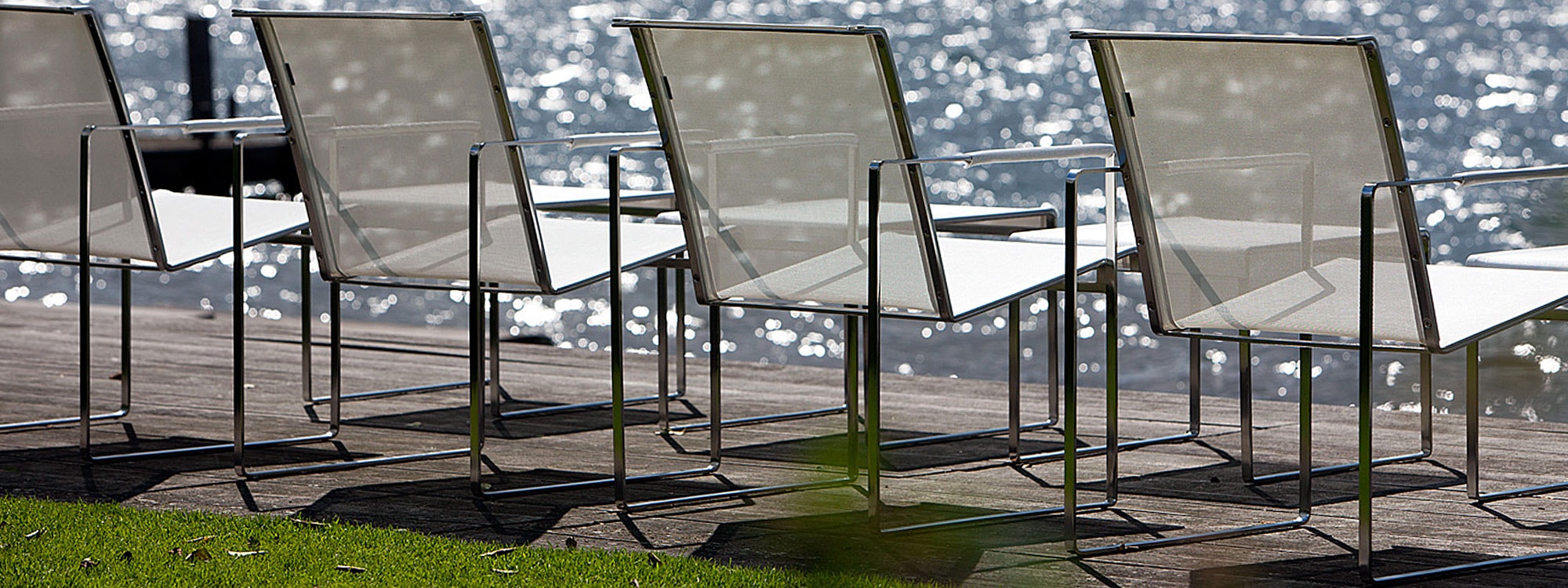 Row of Poltrona outdoor lounge chairs and foot stools designed by Henk Steenbakkers on sunny decking next to Dutch lake