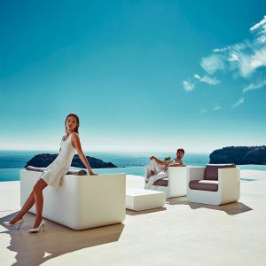 Image of couple leaning of different elements of Vondom Ulm white outdoor lounge furniture on sunny terrace overlooking the blue sea