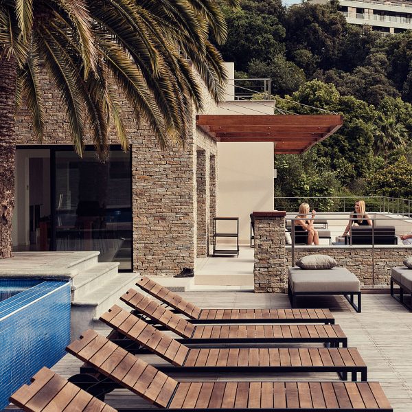 Image of row of Roshults modern sun loungers next to edge of horizon swimming pool, with stone building and fronds of a palm tree in the background