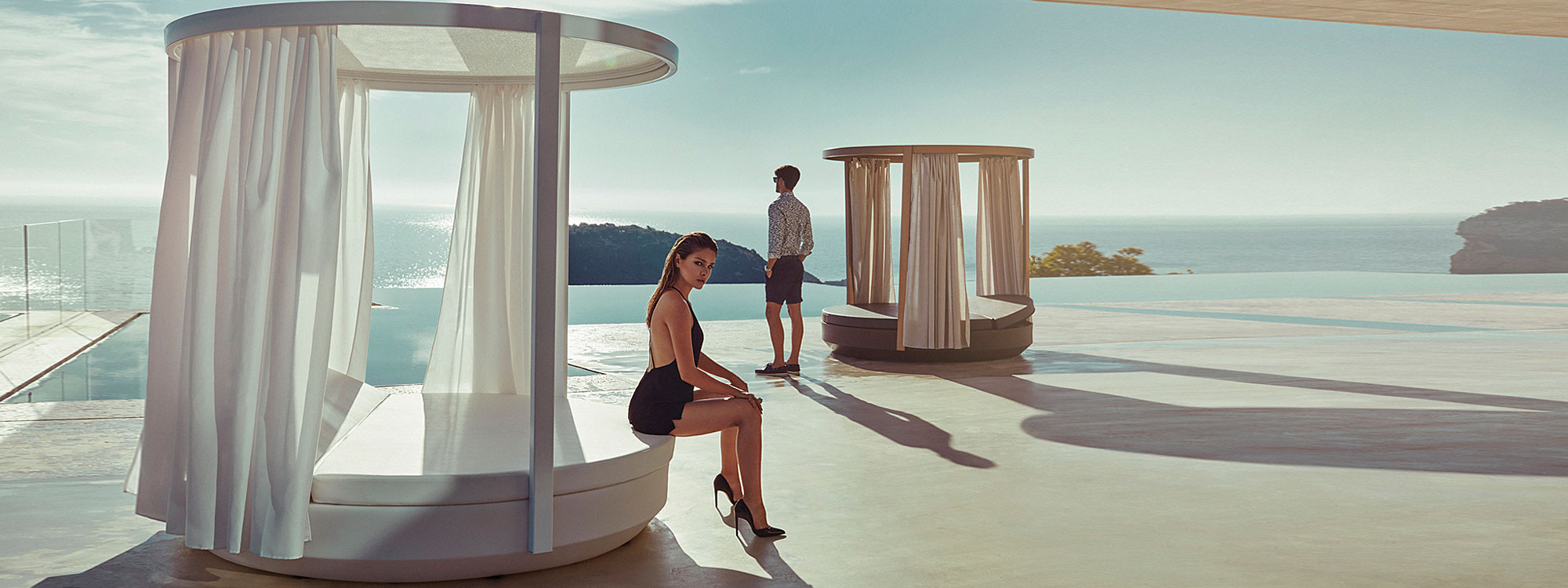 Image of pair of circular Vondom Vela daybeds with integrated pergolas and curtains, shown in late afternoon sun on balcony with hills and sea in the background