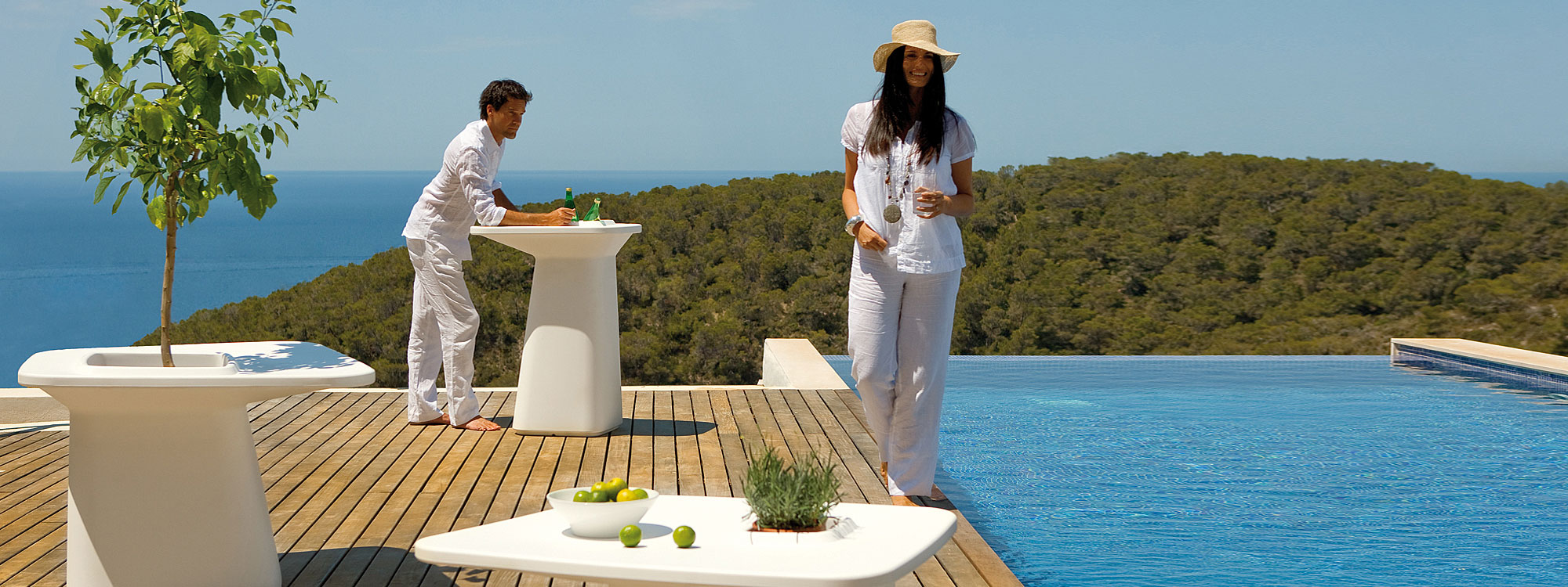 Image of couple dressed in white hanging out around white Moma plastic bar furniture by Vondom on sunny poolside, with tree covered hill, sea and sky in the background