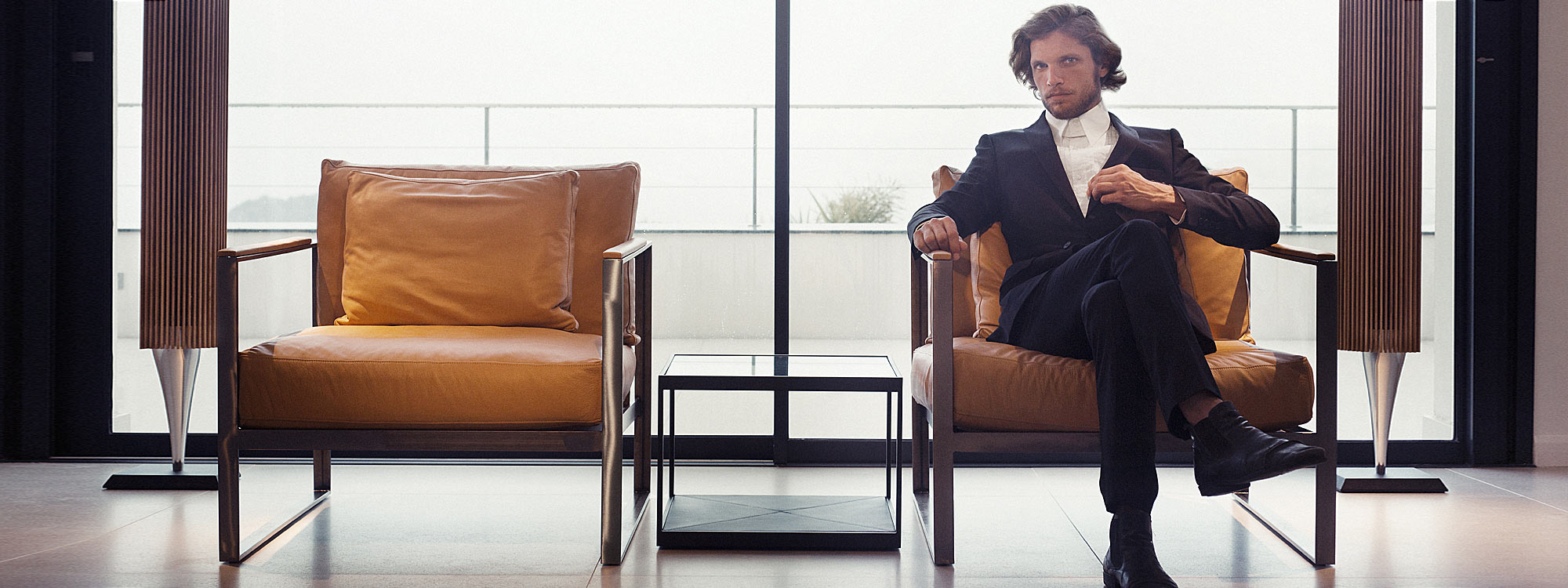 Image of Swedish hipster dressed in a suit, sat in Roshults Monaco lounge chair in Raw stainless steel with Beige leather cushions, by Roshults Sweden