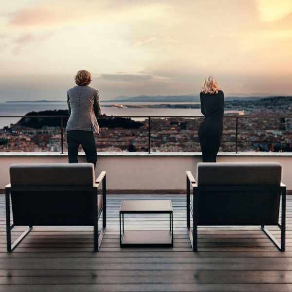 Image of pair of Roshults Garden Easy chairs on terrace at dusk, with couple stood at railings looking out towards distant coastal town and sea