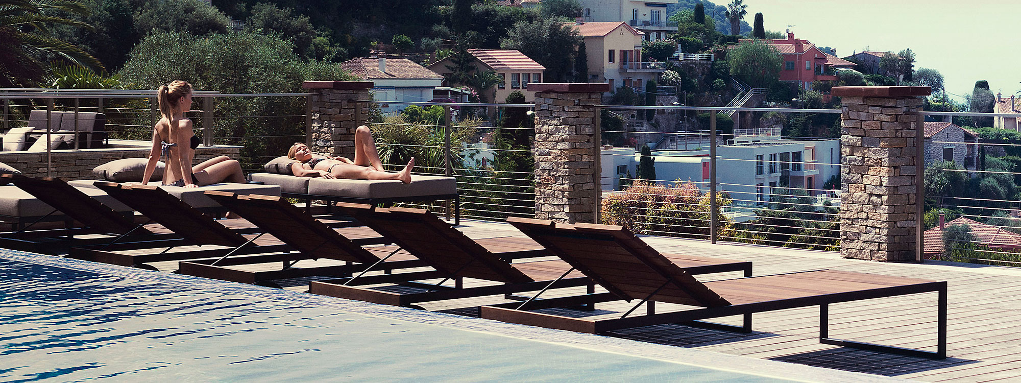 Image of row of Roshults Garden Loungers on poolside next to 2 women relaxing on Garden Easy daybeds, with hillside dotted with villas in the background