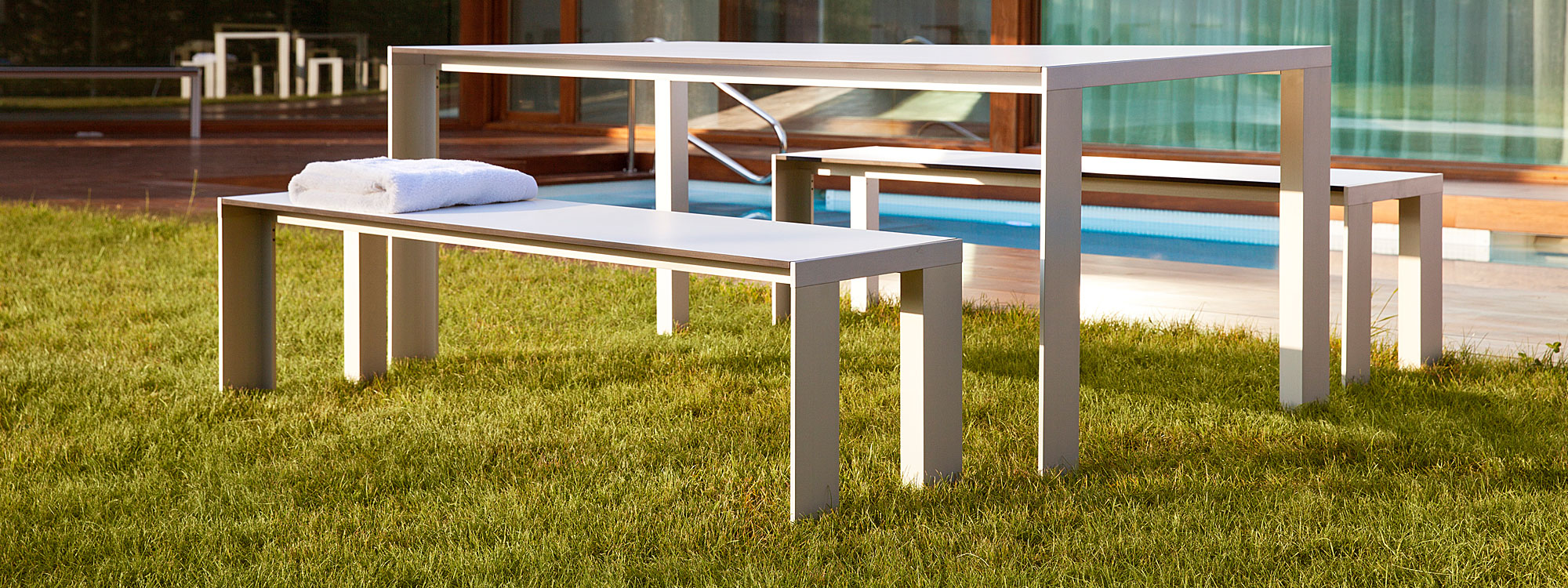 Image of Stua Deneb minimalist white garden table and benches with white HPL surfaces, shown in grassy lawn with swimming pool and building in the background