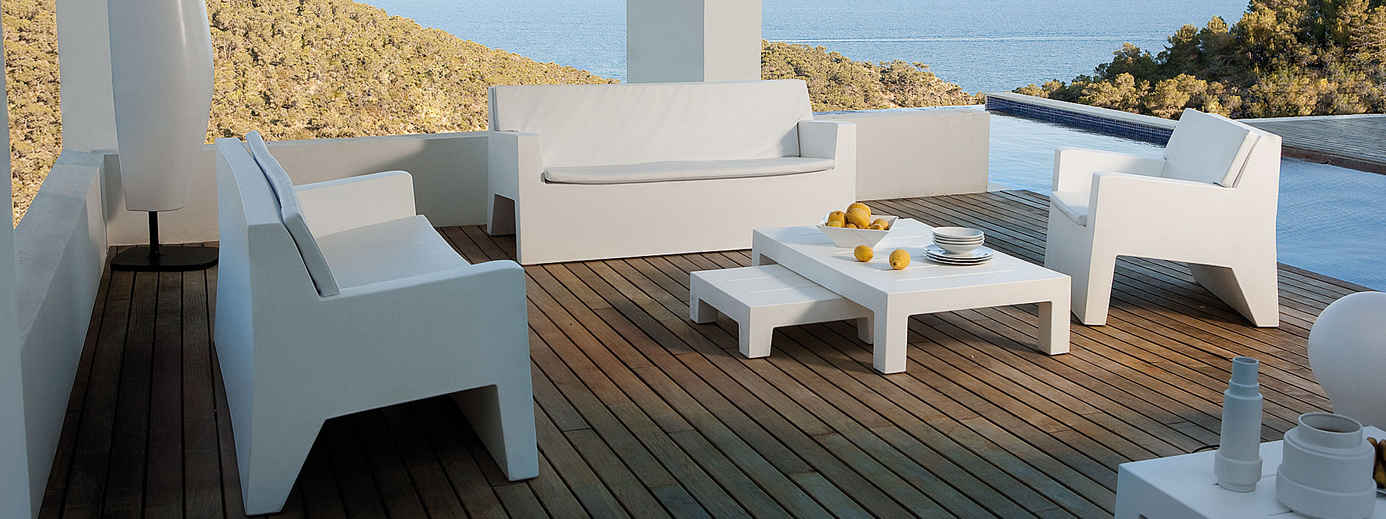 Image of white Jut modern plastic sofa, lounge chairs and low tables by Vondom, on covered pergola, with woodland and sea in the background