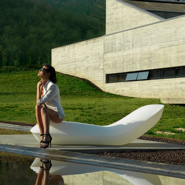 Image of woman sat on Vondom Pillow modern sun lounger beside swimming pool with brutalist architecture and lawn in the background