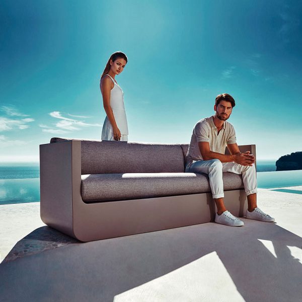 Image of couple stood and sat around Vondom Ulm bronze garden sofa in late afternoon sun, with sea and sky in the background
