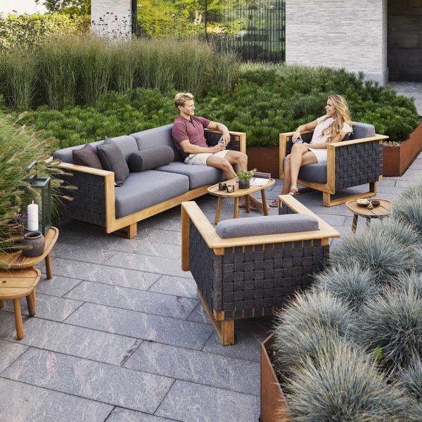 Image of couple sat on Cane-line Angle teak lounge furniture, surrounded by raised beds of grasses