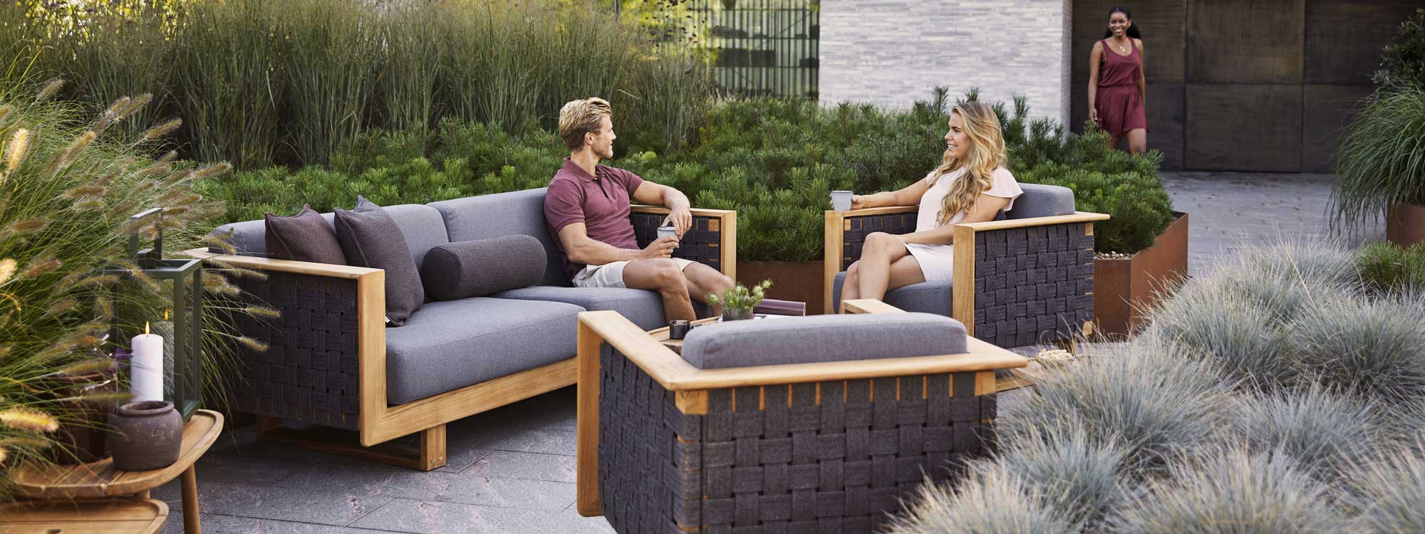 Image of couple relaxing in Caneline Angle teak sofa and lounge chair, surrounded by raised beds of grasses