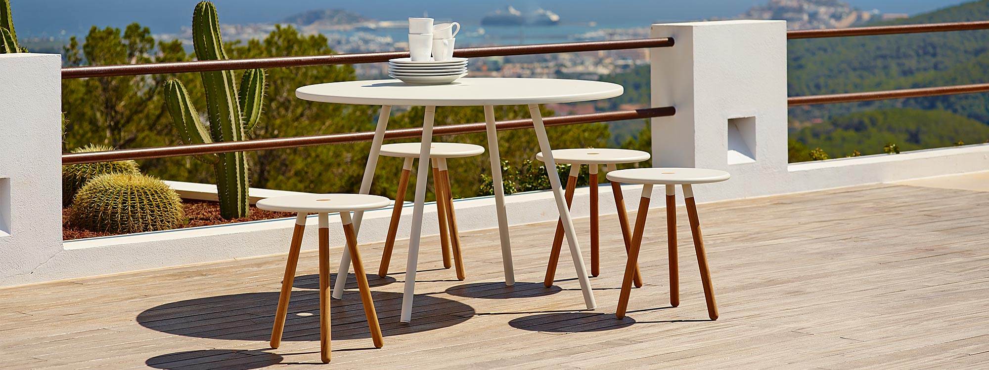 Image of Caneline Area round garden table and stools in white aluminum and teak, shown on terrace with cactuses and port in the background