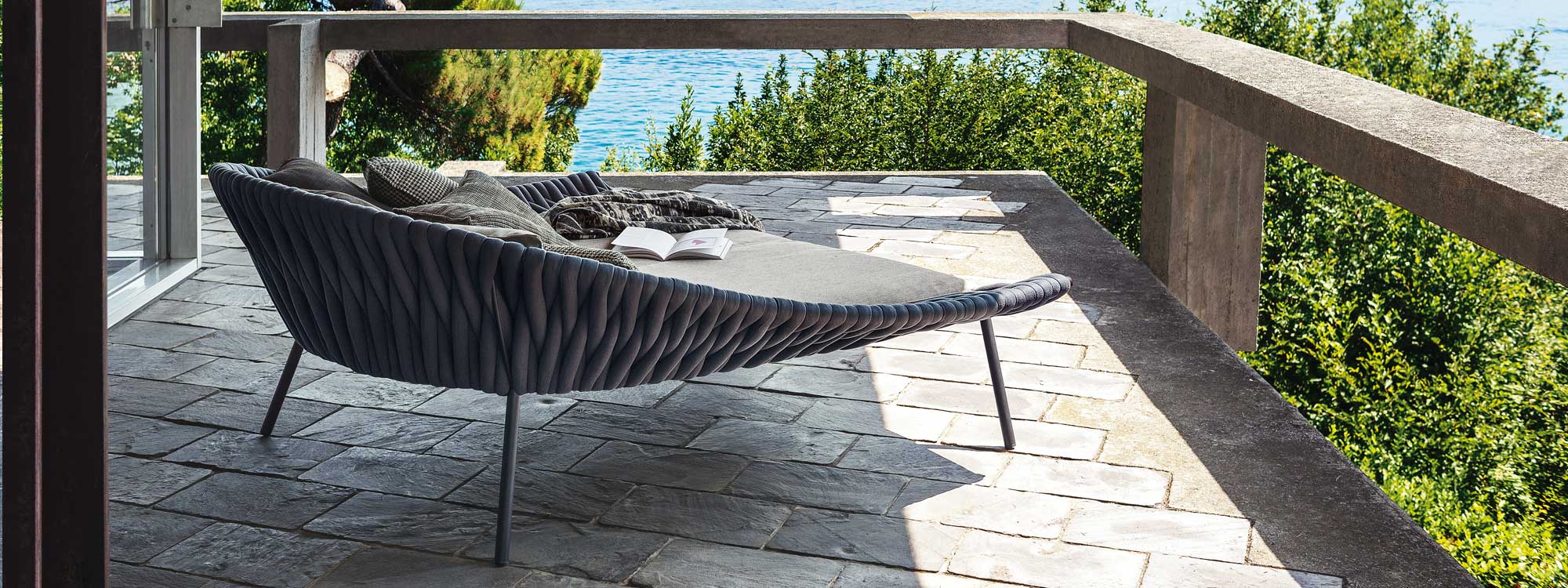 Image of RODA Arena daybed with book and scarf on top of cushions, shown on terrace looking out to sea