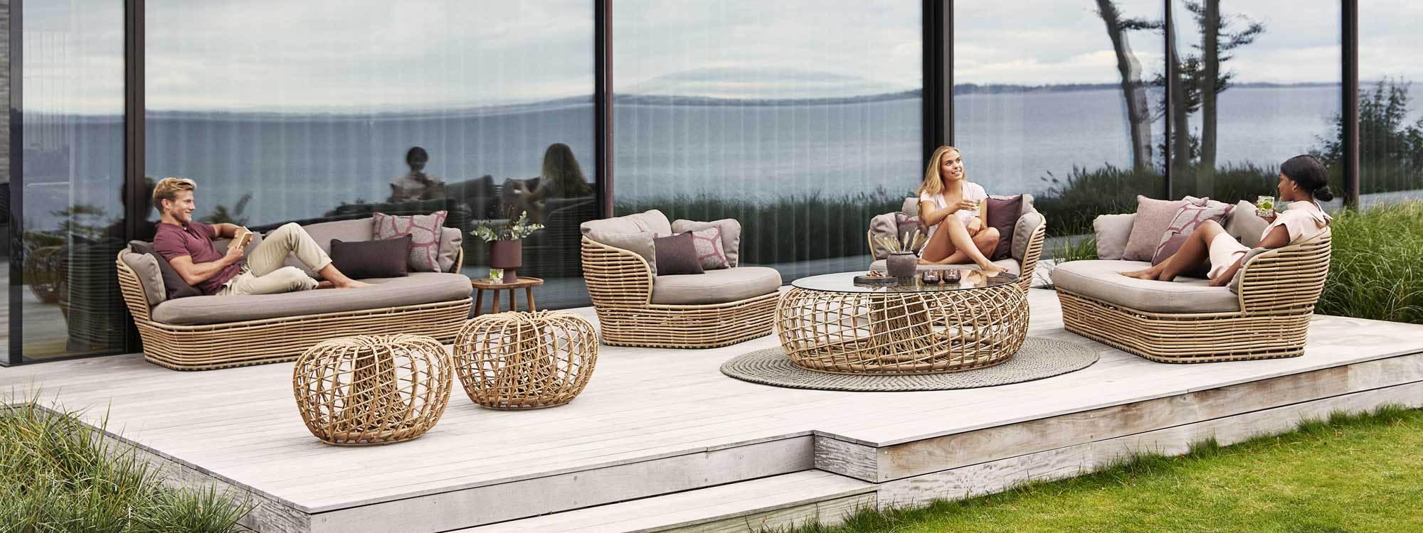 Image of man and 2 women relaxing on Cane-line Basket cane garden sofas on decking, with Nest cane garden tables in the centre