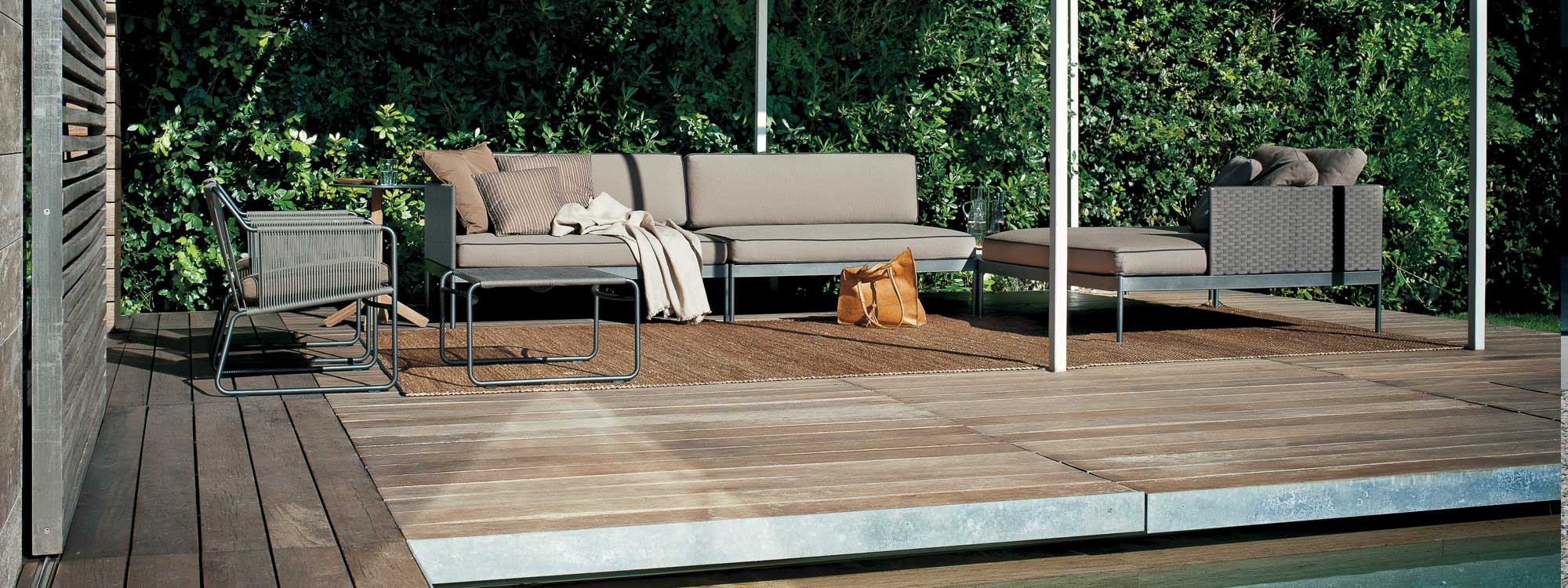 Image of Basket modern outdoor sofa with smoke colored frame and taupe cushions together with Harp lounge chair and foot stool, shown on minimalist wooden deck with plants in background