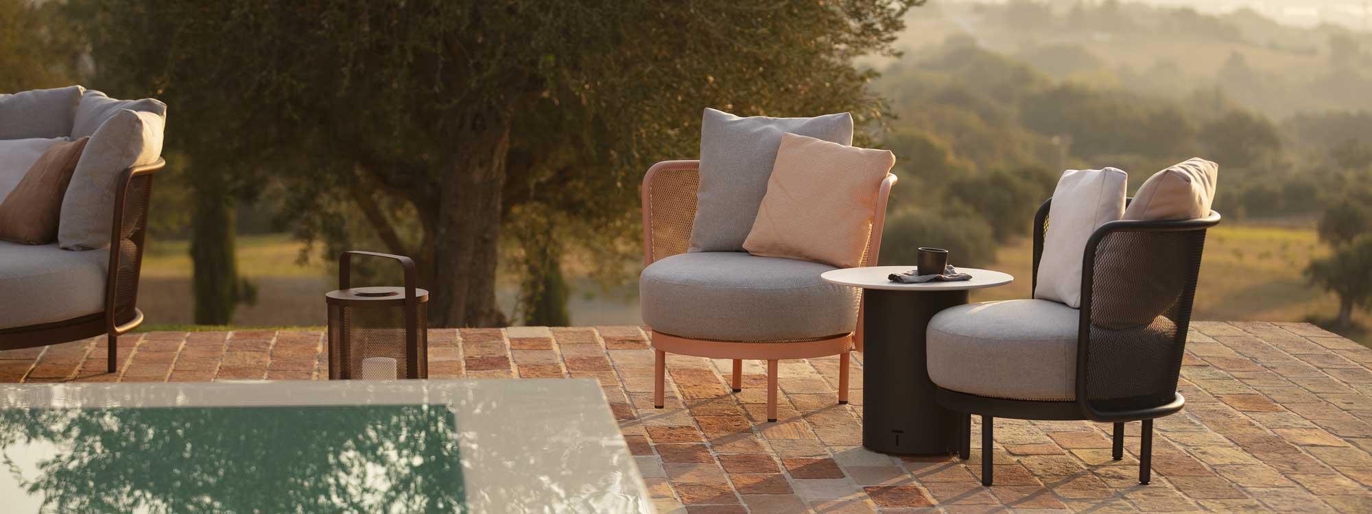Image of salmon-pink and anthracite Baza Club lounge chairs and Branta modern garden side table on sunny poolside, with rolling countryside and woodland in the background