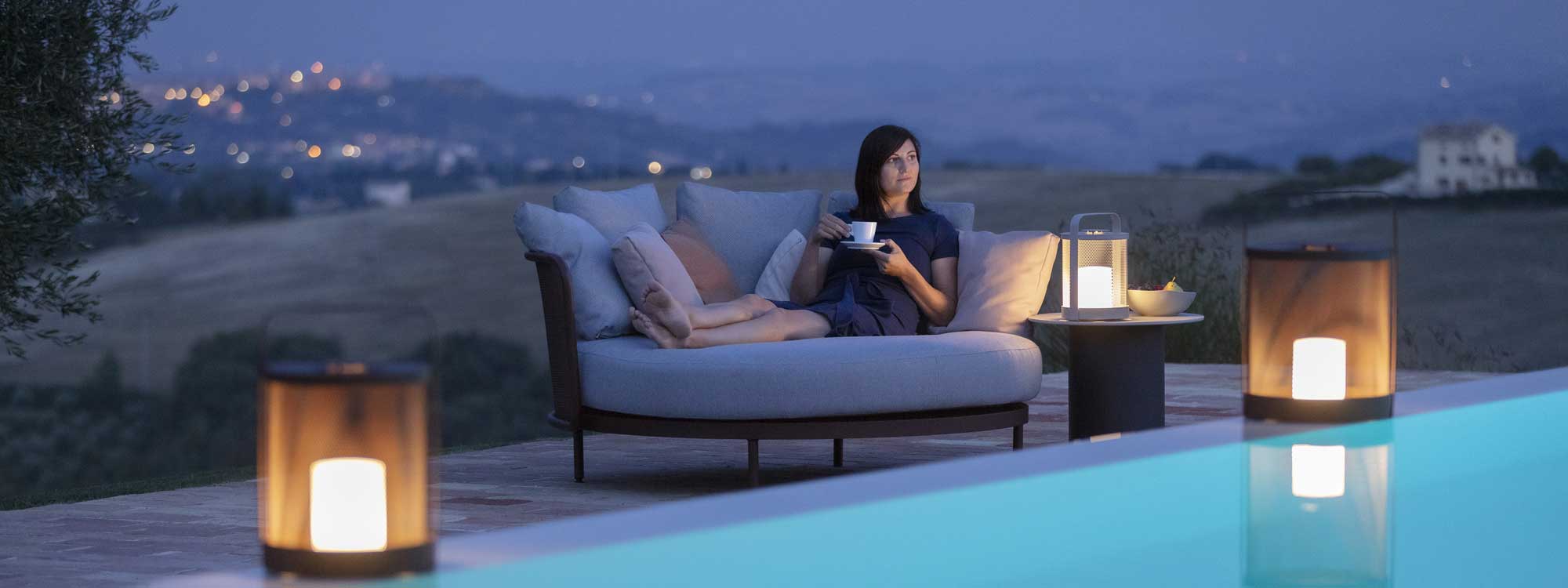 Dusk shot of woman relaxing in Baza modern garden day bed next to swimming pool, with lights of town twinkling in distance