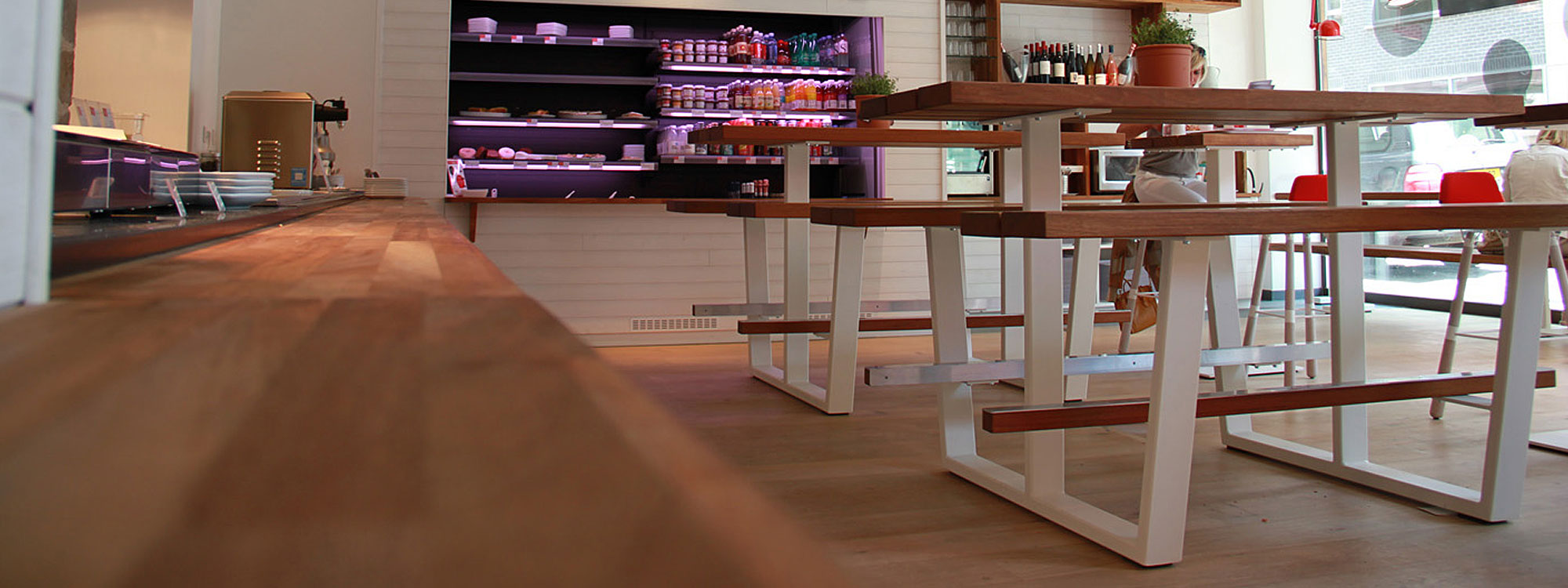 Image of pair of Cassecroute Beer tables with white frames and integrated bench seats, with surfaces in iroko, shown in indoor kitchen