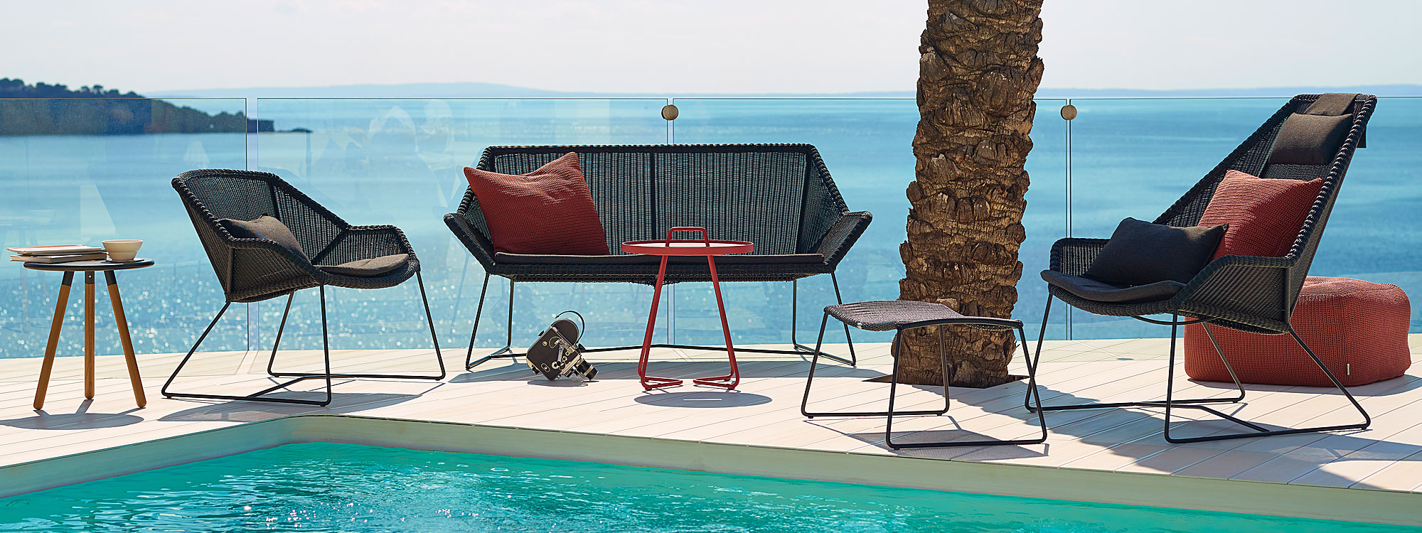 Image of Cane-line Breeze 2 seat sofa and lounge chairs in black Cane-line weave, together with On The Move outdoor side tables on poolside, with palm tree trunk to one side and sparkling sea in the background