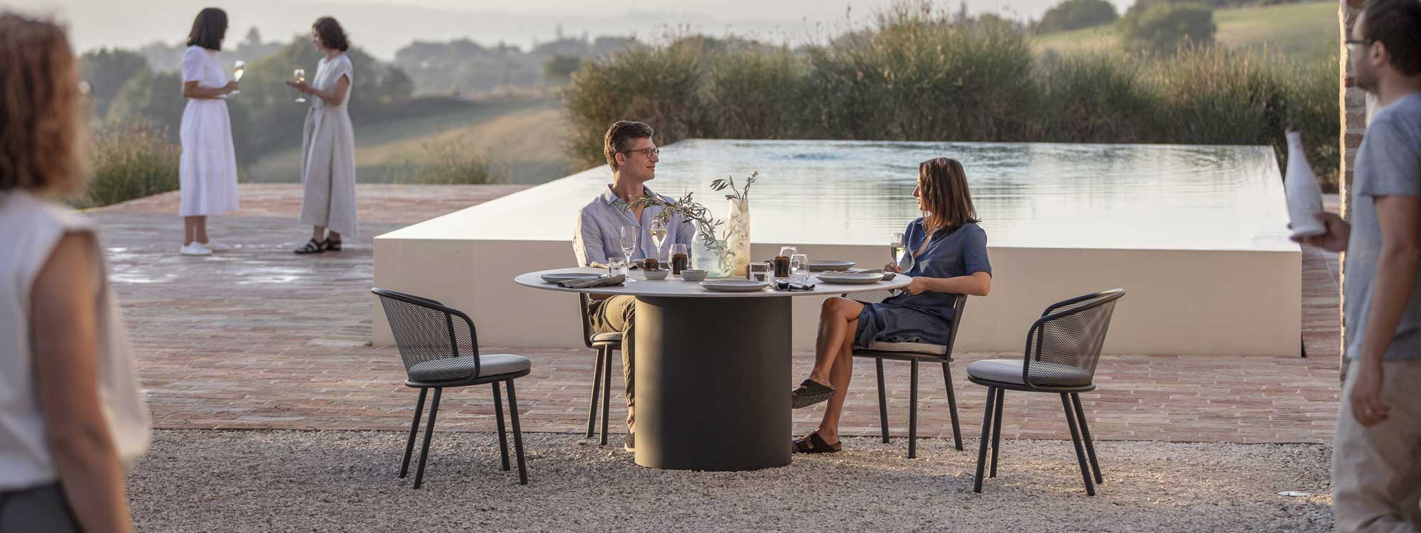 Branta large round garden table and Baza outdoor dining chairs on terrace in front of swimming pool and olive trees