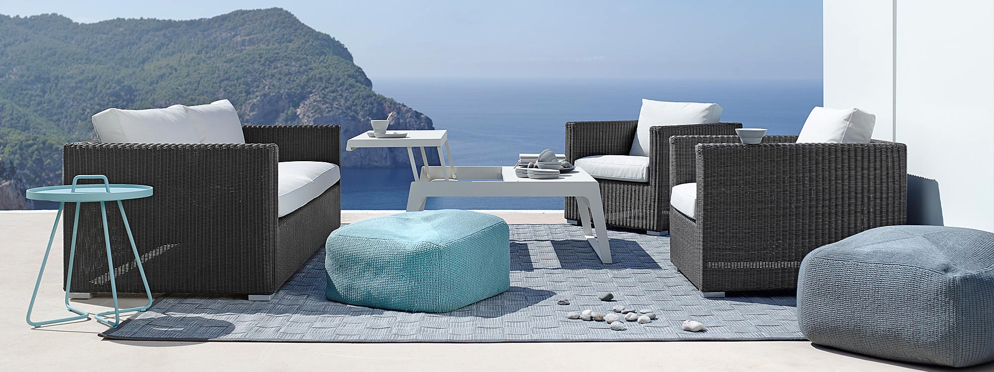 Image of Cane-line Chester black rattan garden sofa, On The Move tray table and Divine outdoor pouf on terrace with sea and cliffs in background