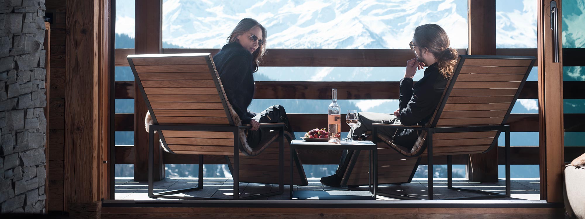Image of pair of couple of hipsters sat in Roshults Garden Sun Chairs on chilly terrace, with snowy mountain peaks in the background