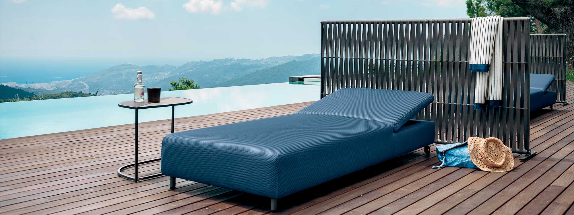 Image of Double minimalist sun lounger upholstered in blue 3d fabric, with Wing outdoor screen and horizon swimming pool in the background
