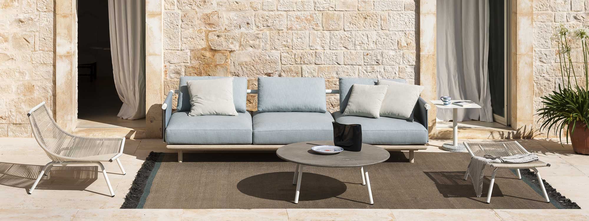 Image of Eden modern garden sofa with oiled white teak base and light blue cushions, with Laze minimalist low chair and foot rest and Piper stone low table by RODA