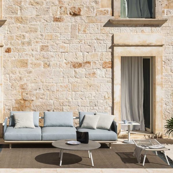 Image of Eden modern teak garden sofa with light blue cushions, shown on sunny terrace outside rustic building